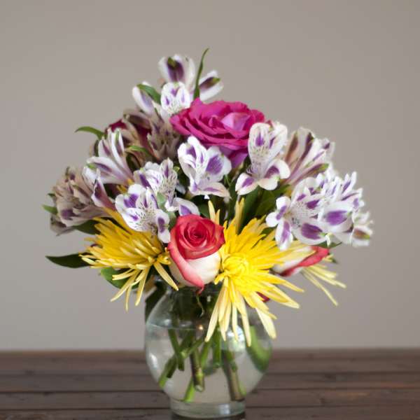 Mixed bouquet of pink roses, yellow spider mums, and white-purple alstroemeria in a glass vase