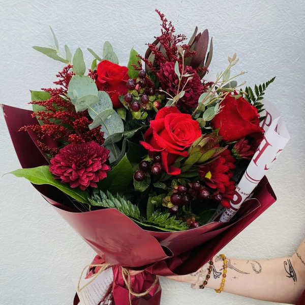 Hand-tied bouquet with red roses, red chrysanthemums, and berries wrapped in dark red paper.