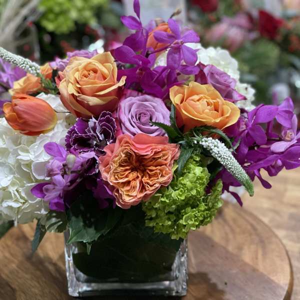 Bright mixed arrangement of orange roses, purple orchids, and hydrangeas in a glass cube vase