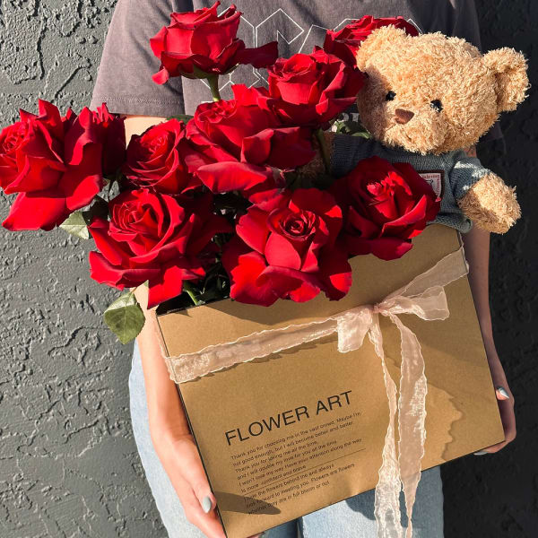 Box of red roses with a small teddy bear held in someone’s arms