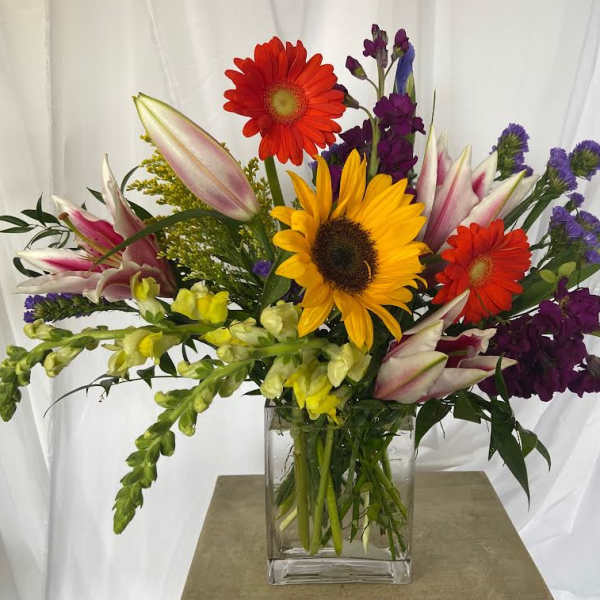 Mixed bouquet with sunflowers, gerbera daisies, lilies, and purple blooms in a glass vase