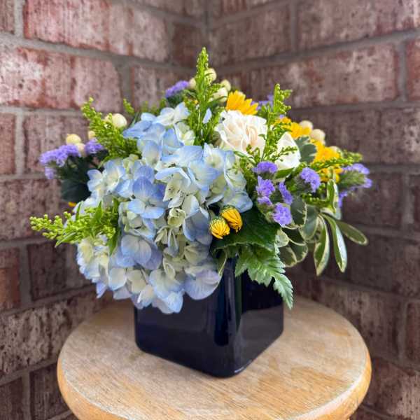 Blue hydrangea bouquet in a black square vase with yellow and purple accents
