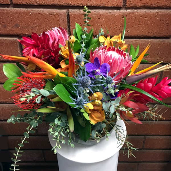 Vibrant tropical arrangement with protea, orchids, and birds of paradise in a glass vase on a white stand