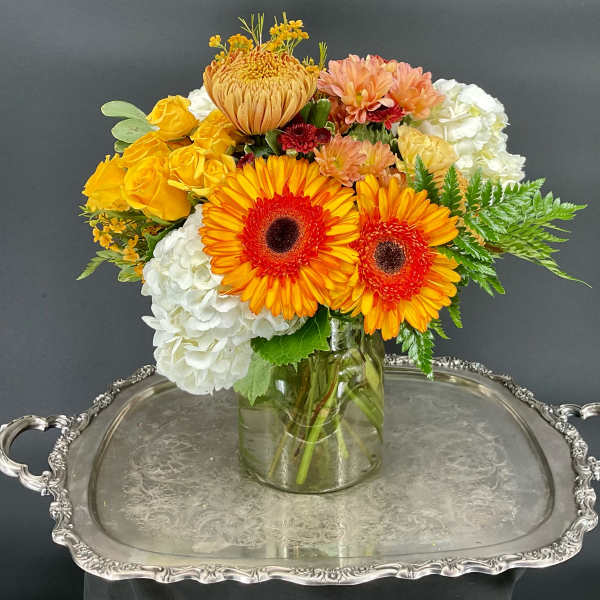 Bright mixed bouquet in a glass vase with orange gerberas, yellow roses, and white hydrangeas.