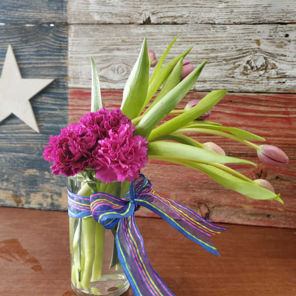 Tall glass vase with bright pink carnations and closed pink tulips, tied with a striped ribbon.