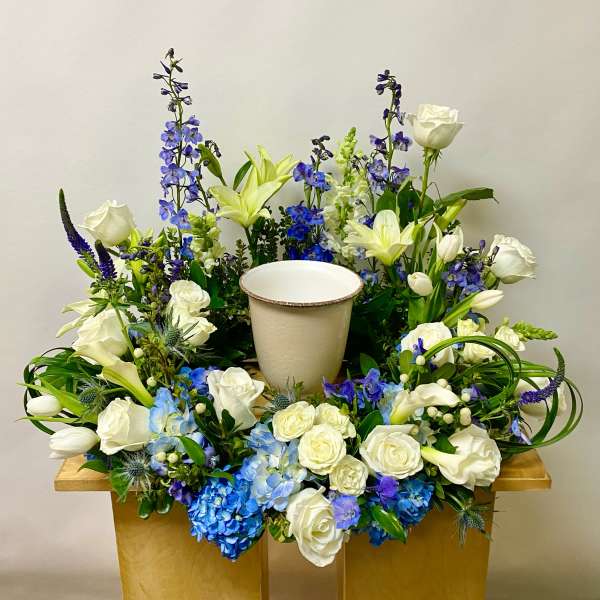 White roses and blue flowers arranged around a central urn on a wooden stand.