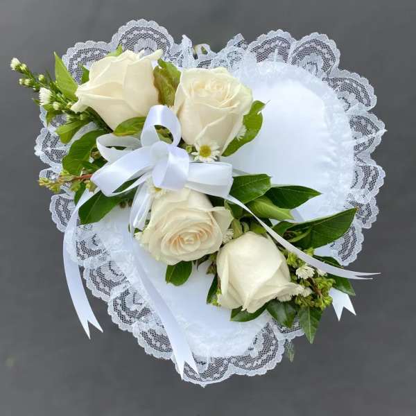 White heart-shaped lace pillow adorned with ivory roses and a white ribbon bow.