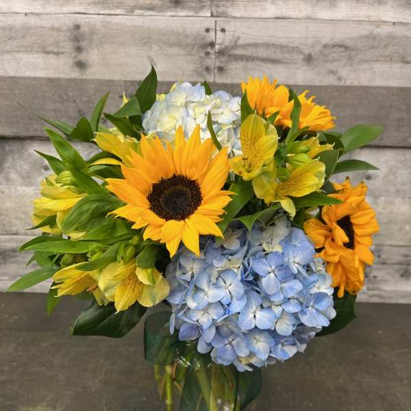 Bouquet of sunflowers, blue hydrangeas, and yellow alstroemeria in a glass vase