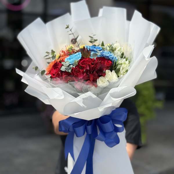 Bouquet of red, blue, white, and orange flowers wrapped in white paper with a blue ribbon