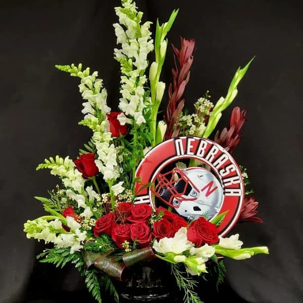 Red roses and white snapdragons arranged with a Nebraska football emblem.
