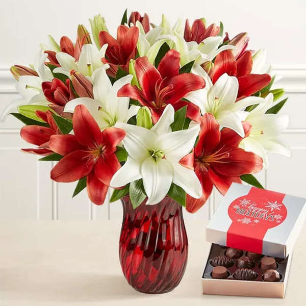 Red and white lilies in a red glass vase beside an open box of chocolates