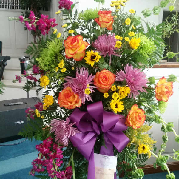 Tall floral standing spray with orange roses, purple mums, and yellow daisies