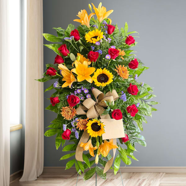 Standing floral spray with red roses, sunflowers, and orange lilies on a ribboned easel