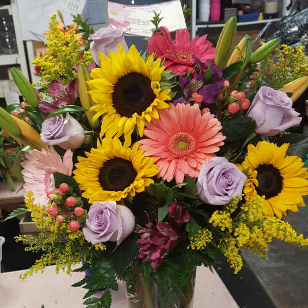 Mixed bouquet with sunflowers, gerbera daisies, and lavender roses in a glass vase
