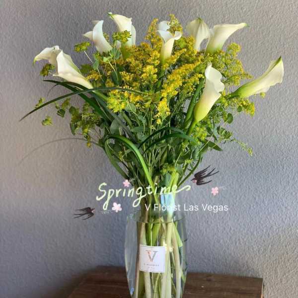 White calla lilies and yellow filler flowers in a clear glass vase
