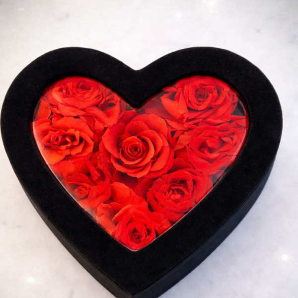 Heart-shaped black box filled with red roses under a clear lid on a light surface