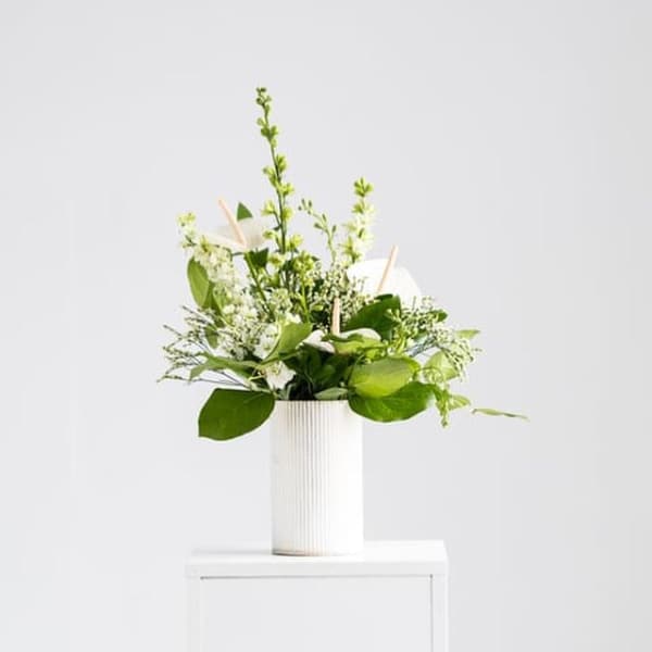 White and green floral arrangement in a tall white ribbed vase on a white pedestal