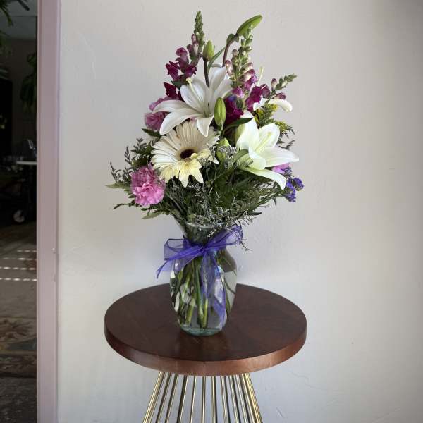 Tall mixed bouquet of white lilies and daisies with purple and pink flowers in a clear vase with a purple bow.