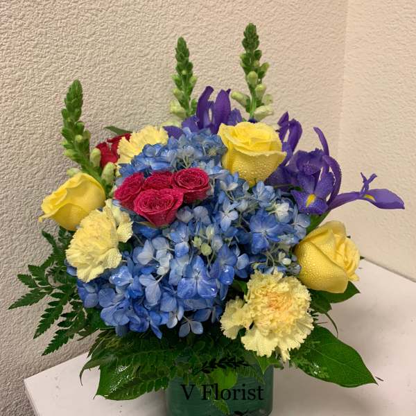 Bouquet of blue hydrangeas, yellow roses, and purple irises in a square vase
