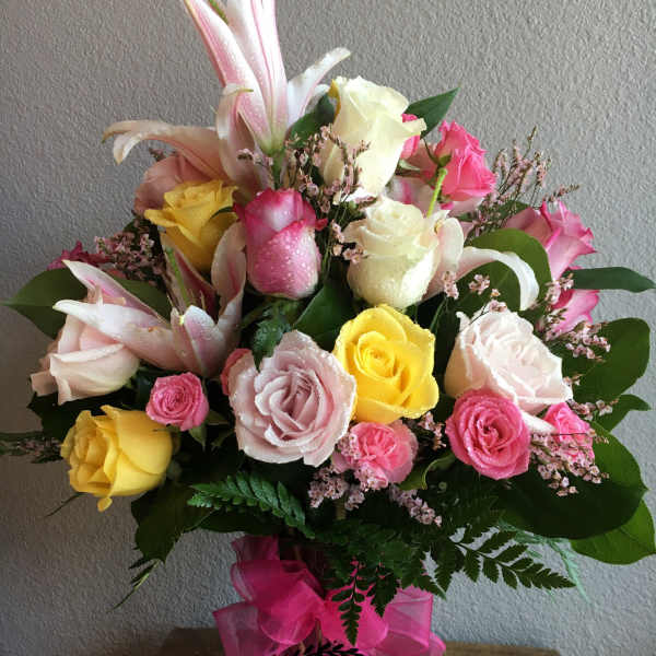 Tall mixed arrangement of pink lilies and pink, yellow, and white roses in a clear vase with pink bow