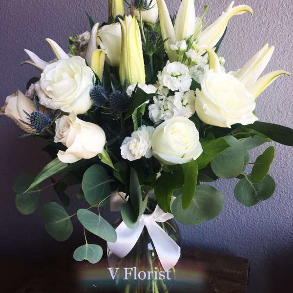 Tall arrangement of white lilies and roses with small white blooms in a clear glass vase with a white bow