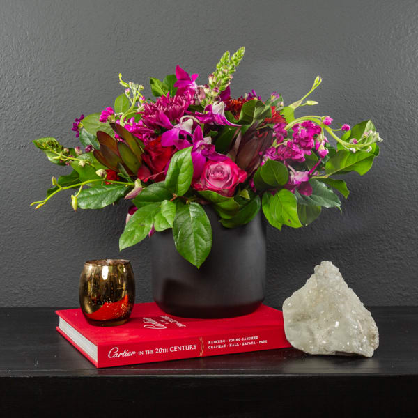 Pink and purple flower arrangement in a black vase with a gold candle and crystal.