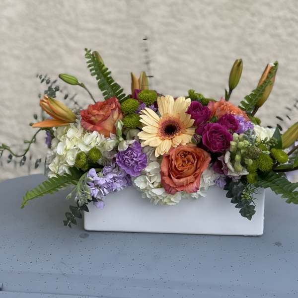 Mixed floral arrangement in a white rectangular vase with peach, purple, and white blooms