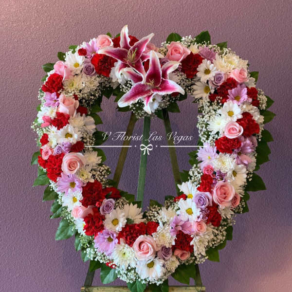 Heart-shaped standing wreath of pink lilies, roses, red carnations, and white daisies on an easel.