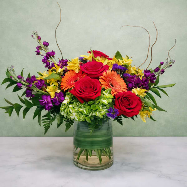 Mixed bouquet of red roses, orange gerberas, and purple flowers in a glass vase