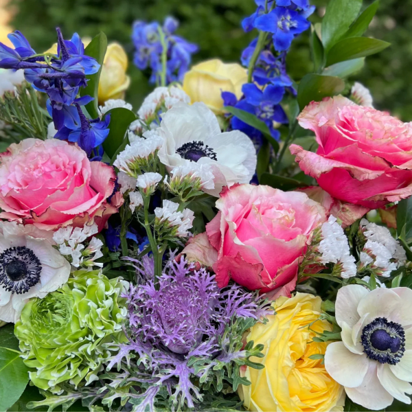Colorful arrangement with pink roses, white anemones, blue delphinium, and other mixed blooms.