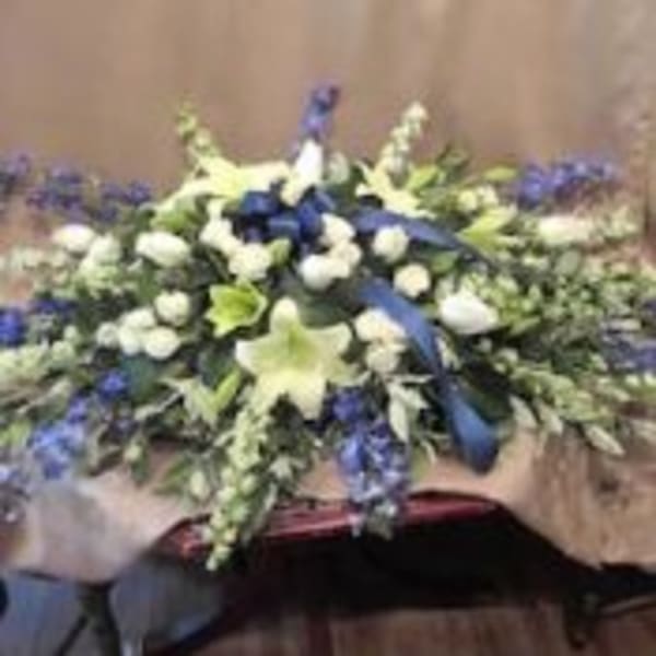 Elongated white and blue funeral spray with lilies, roses, and delphinium accented by a blue ribbon