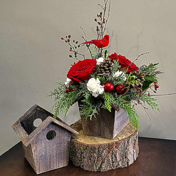 Rustic birdhouse arrangement with red roses, white flowers, pinecones and a red cardinal on a wood slice base.