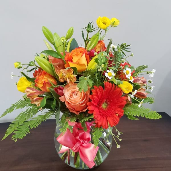 Bright mixed bouquet of orange roses, red gerbera daisy and yellow blooms in a round glass vase with pink bow