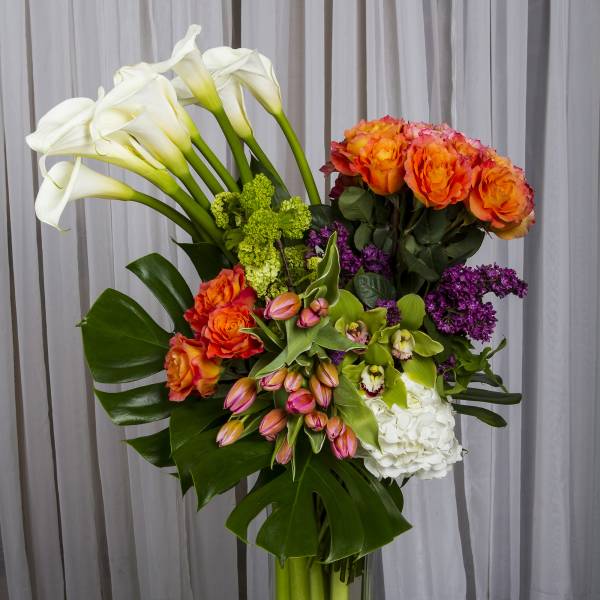 Tall bouquet of white calla lilies, orange roses, and purple flowers in a glass vase