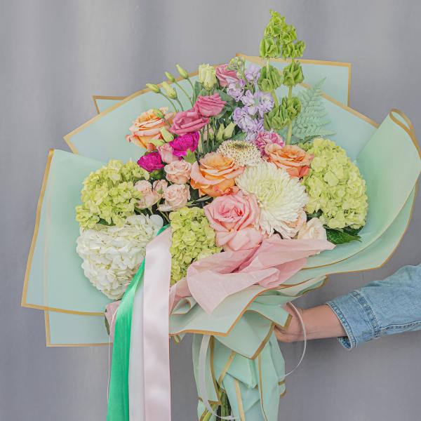 Handheld bouquet of pastel roses, hydrangeas, and chrysanthemums wrapped in mint paper