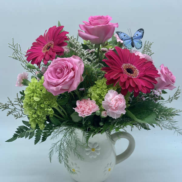 Pink roses, gerbera daisies, green hydrangea, and carnations in a white daisy pitcher with a blue butterfly pick.