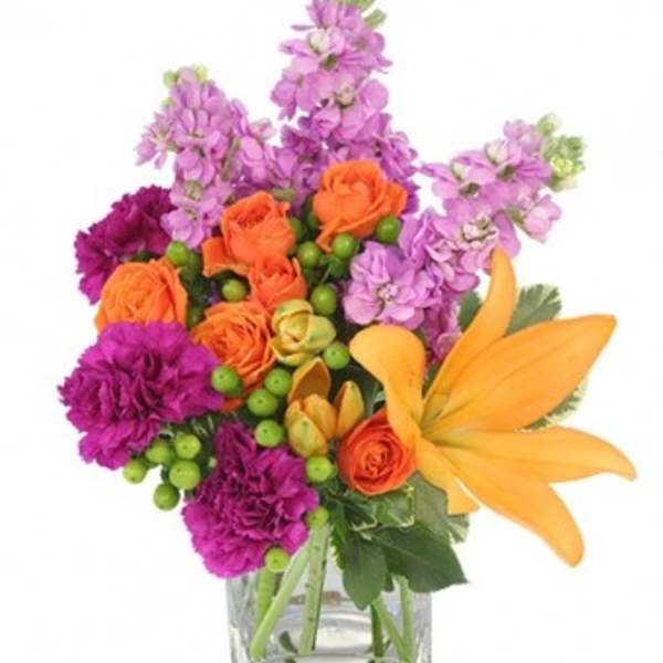 Bright arrangement of orange lilies and roses with purple flowers in a clear square vase