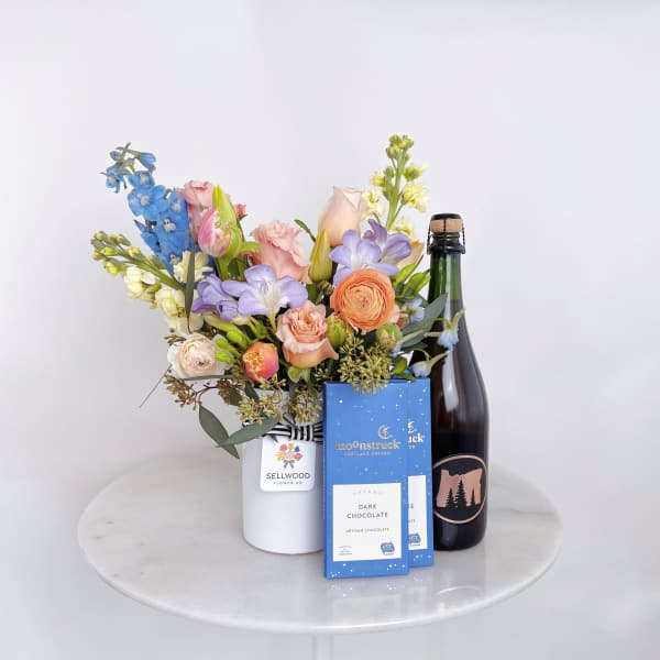 Mixed pastel bouquet in a white vase with chocolate boxes and a bottle of sparkling wine