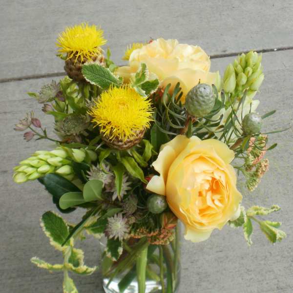 Yellow roses and spiky yellow blooms with green pods in a clear glass vase