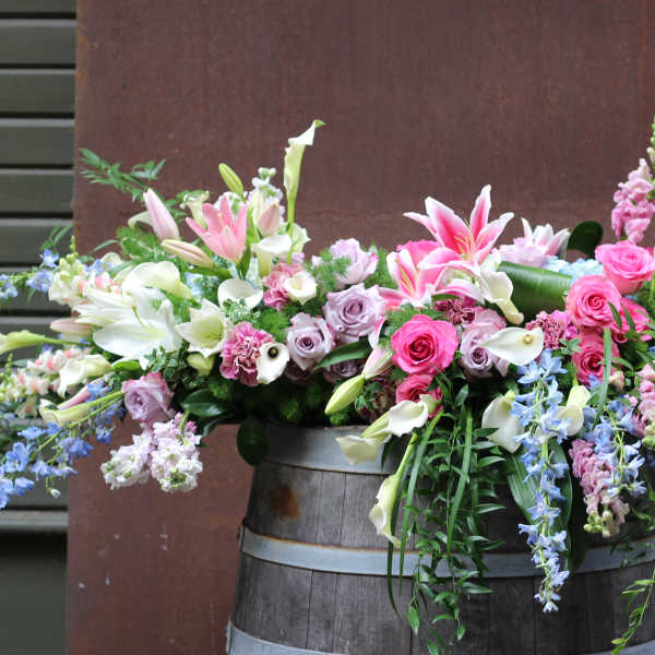 Large mixed floral arrangement with lilies, roses, calla lilies, and snapdragons in a wooden barrel