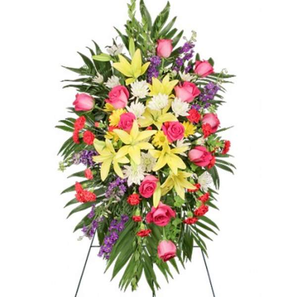 Standing spray of yellow lilies, pink roses, red carnations and purple flowers on lush green foliage.
