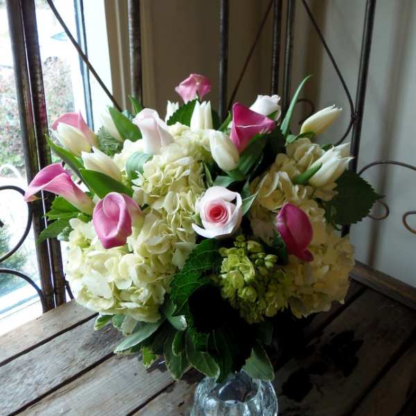 Pink calla lilies and white tulips arranged with pale hydrangeas in a glass vase.
