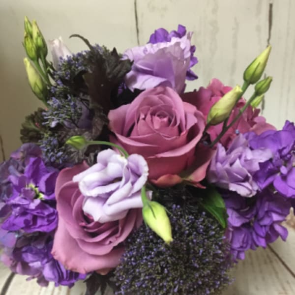 Compact arrangement of purple and lavender roses and blooms in a clear glass cylinder vase