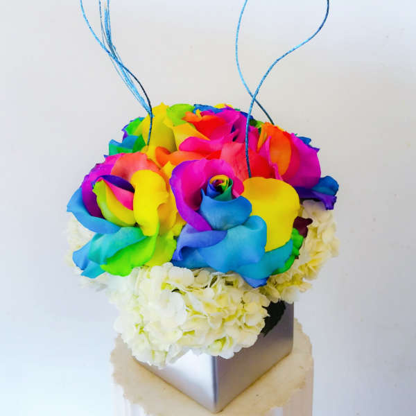 Rainbow-colored roses arranged in a square vase with blue curly picks
