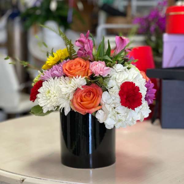 Colorful mixed arrangement with orange roses, white blooms, and red accents in a black cylinder vase