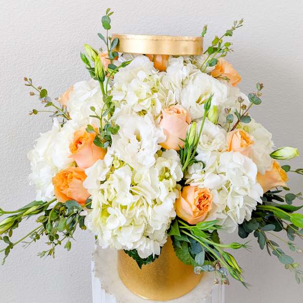 White and peach floral arrangement in a gold hatbox-style container
