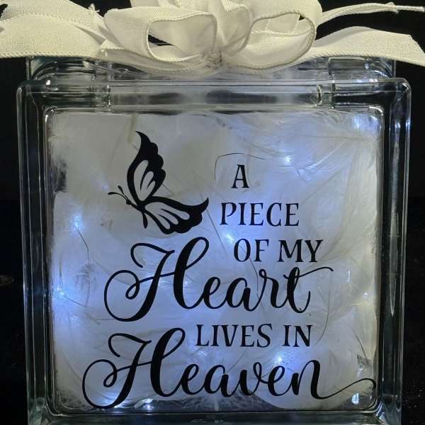 Glass block light with white bow, feathers, and memorial text 'A piece of my heart lives in Heaven'.