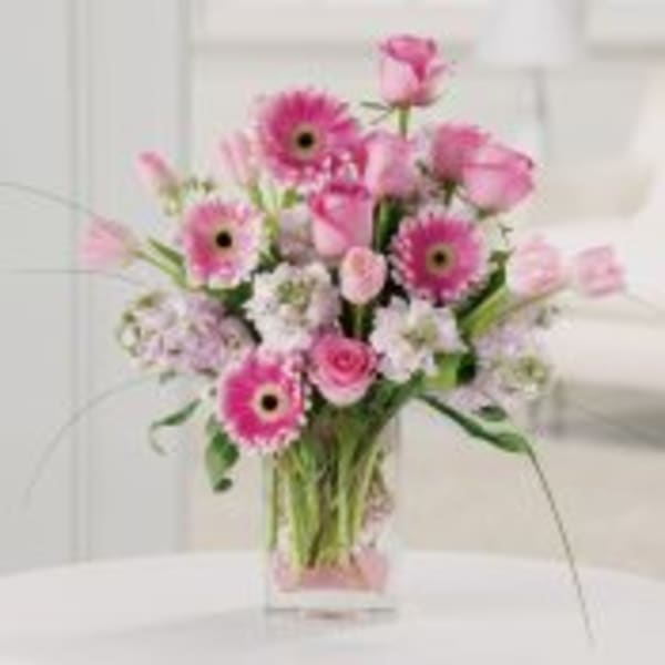 Pink gerbera daisies, roses, and other blooms arranged in a clear rectangular vase.