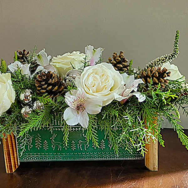 Low rectangular holiday arrangement with white flowers, pinecones, and evergreens in a green wooden box.