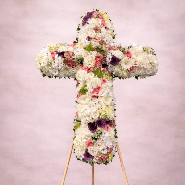 Standing floral cross of white blooms with pink roses and purple accents on a wooden easel